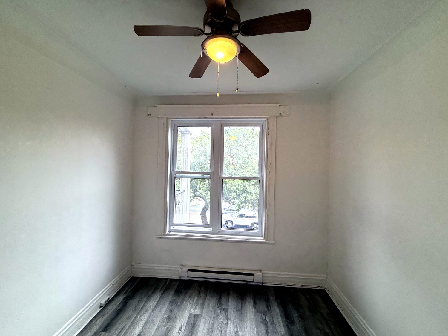 Petite chambre vide avec fenêtre sur la rue et ventilateur de plafond.