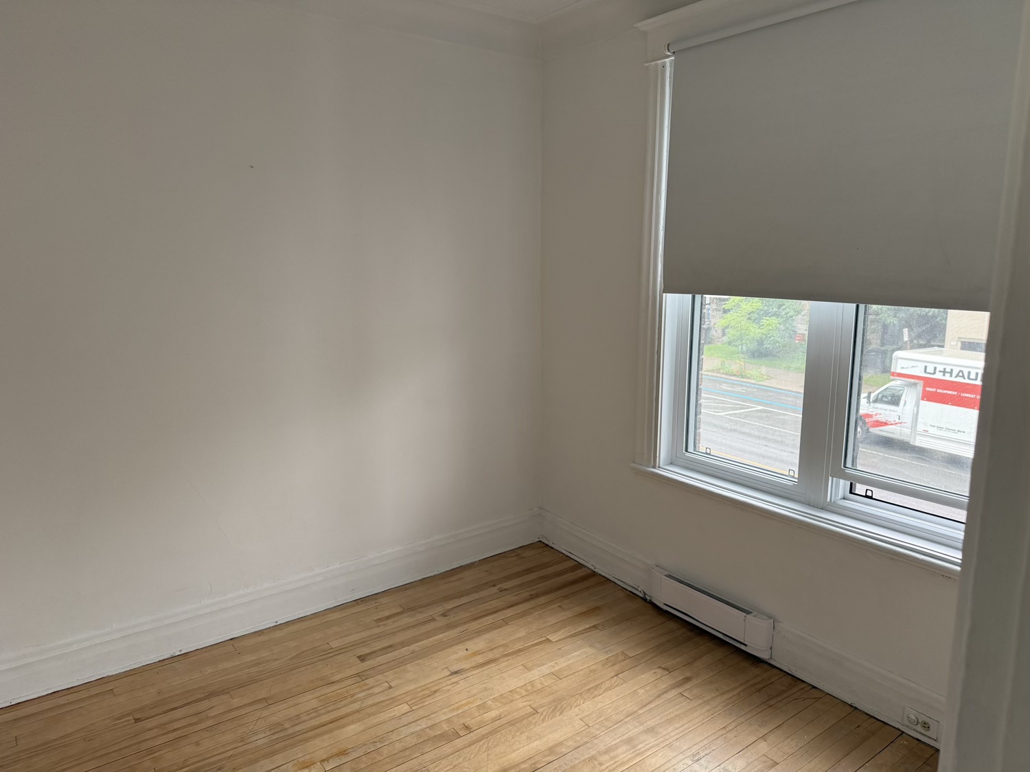 Chambre vide avec store, grande fenêtre et plancher de bois clair.